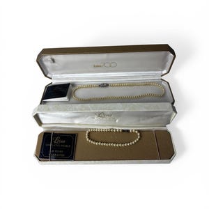 May include: A jewellery box containing a pearl necklace and bracelet. The necklace and bracelet are composed of small, round, off-white pearls. The box has a gold-coloured exterior and is lined with white satin. The brand name "Lotus" is visible.