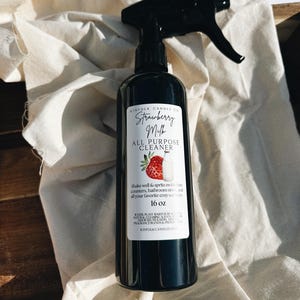 May include: A black spray bottle of "Strawberry Milk All Purpose Cleaner" by Kinfolk Candle Co. The label features a strawberry and milk bottle illustration. The bottle contains 16 oz of cleaner and is plant-based.