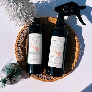 May include: Two black bottles of cleaning products, "Sugar Plum Fairy" mop soap and all-purpose cleaner, sit in a woven basket. The bottles have white labels with text and a pink illustration. Each bottle is 473 ml.