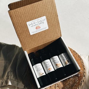 Può includere: Una scatola di cartone marrone contenente quattro flaconi neri di campioni di sapone per mop profumati. Ogni flacone ha un'etichetta bianca con il nome della fragranza e una piccola grafica. La scatola ha un adesivo che dice "Smells like a Sunday reset and emotional stability."