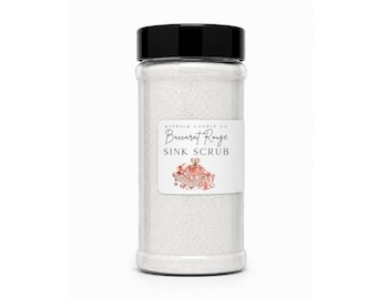 May include: A clear plastic cylindrical container filled with white granular sink scrub. The black screw-top lid sits above a white label featuring the text Kinfolk Candle Co, Baccarat Rouge, and SINK SCRUB, accompanied by a small pink perfume bottle graphic.