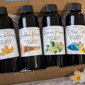 Può includere: Quattro bottiglie nere di campioni di sapone Mop in una scatola di cartone. Ogni bottiglia ha un'etichetta con un profumo diverso: Melon & Honey, Golden Hour, Summer Morning e Citrus Tide. La scatola è su una superficie sabbiosa con fiori e un cappello sullo sfondo.