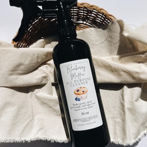 May include: A black spray bottle of "Blueberry Muffin All Purpose Cleaner" with a blueberry muffin graphic. The bottle is 473 ml and sits on a beige cloth with a wicker basket in the background.