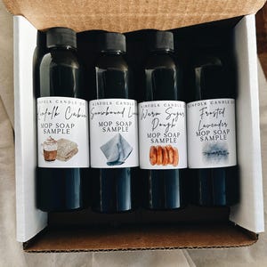 Può includere: Una scatola contenente quattro flaconi neri di campioni di sapone MOP. Ogni flacone ha un'etichetta bianca con testo e un'illustrazione ad acquerello. I profumi sono "Kinfalk Cabin", "Snowbound Lin", "Warm Sugar Dough" e "Frosted Lavender".