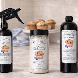 Puede incluir: Tres botellas negras de productos de limpieza "Blueberry Muffin" con ilustraciones de magdalenas y arándanos. Los productos incluyen limpiador multiusos, ambientador para alfombras y jabón para fregar, cada uno de 473 ml.