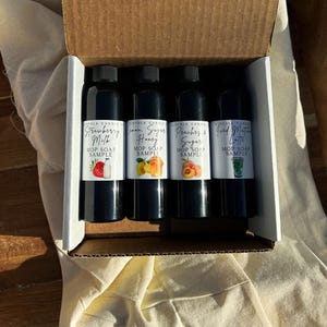 Può includere: Una scatola di cartone contiene quattro bottiglie nere di Mop Soap Sample. Le etichette riportano Strawberry Milk, Lemon, Sugar, Honey, Peaches & Sugar e Good Matcha Latte. Ogni etichetta ha un'illustrazione di frutta o bevanda.