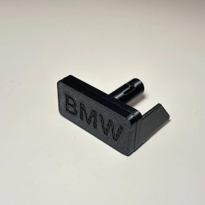 以下が含まれることがあります： 長方形のベースに「BMW」の文字がエンボス加工された黒色の3Dプリントパーツ。円筒形の突起と直角の延長部があります。これはおそらく自動車部品またはアクセサリーです。
