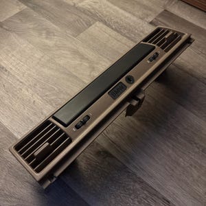 May include: A brown car interior air vent with a black center panel and control dials. The vent has a rectangular shape with a BMW logo.