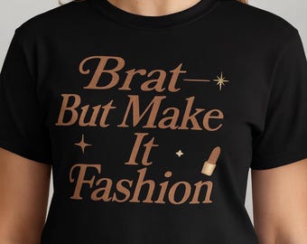 May include: Black t-shirt with the phrase "Brat - But Make It Fashion" in a copper-colored font. The design includes a star and lipstick graphic. The shirt is a crew neck style.