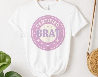 May include: White t-shirt with a round graphic that says "CERTIFIED BRAT" in a purple and cream color scheme. The graphic is surrounded by a pink circle and star accents. The shirt is hanging on a wooden hanger.