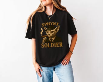 May include: Black t-shirt featuring a graphic of a Sphinx cat wearing a hat and suit, holding a cigar. The words "SPHYNX SOLDIER" are printed in gold lettering above and below the cat illustration.