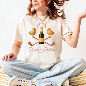 May include: Cream-colored t-shirt featuring a golf-themed design. The graphic includes two yellow birds, a champagne bottle, and crossed golf clubs. The text "Birdies & Bubbles Country Club" is also displayed.