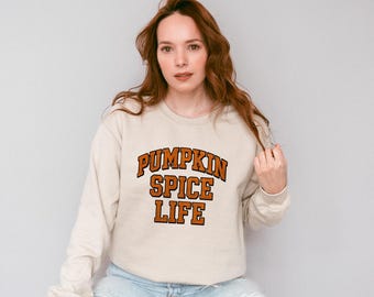 May include: A beige sweatshirt with the text "PUMPKIN SPICE LIFE" in orange and brown lettering. The sweatshirt is a crew neck style and is worn by a person. The overall theme is autumn and fall.