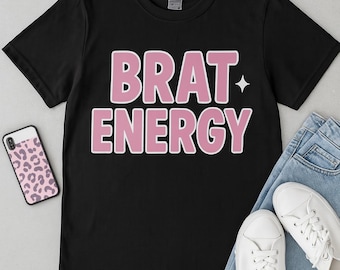 May include: Black t-shirt with the words "BRAT ENERGY" in pink bubble letters. A small white star is next to the word "ENERGY". The shirt is paired with blue jeans, white sneakers, a phone case with a pink leopard print, and round glasses.