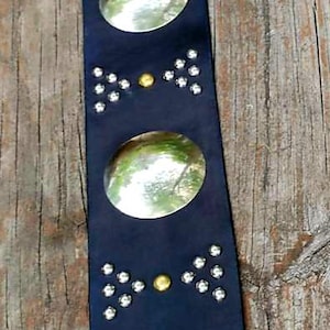 May include: A dark blue leather belt with large, oval, silver-colored embellishments. The belt also features smaller silver and gold-colored studs. The belt is shown against a wooden background.