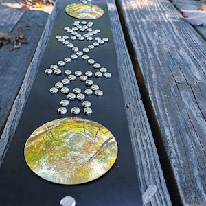 May include: A black leather belt with silver-colored studs arranged in a geometric pattern. Two large, gold-colored circular embellishments are present. The belt is displayed on a weathered wooden surface, suggesting an outdoor setting.