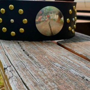 May include: A black leather cuff bracelet with gold and silver studs and two large silver mirrored circles. The bracelet is resting on a weathered wooden surface.