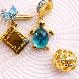 Op de afbeelding: Gouden bedels met blauwe accenten. De bedels omvatten een schildpad met een blauwe edelsteenlichaam, een boek, een libel en een bolvormige bedel met een honingraatontwerp. De bedels worden weergegeven op een wit, getextureerd oppervlak.