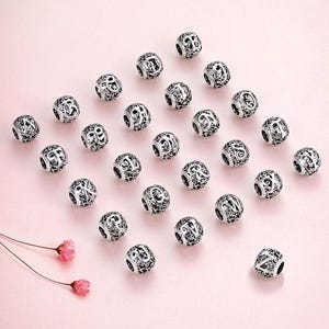 Op de afbeelding: Zilverkleurige alfabetkralen met ingewikkelde openwerkontwerpen liggen op een roze ondergrond. Elke kraal heeft een andere letter, met een bloemenpatroon. De kralen zijn rond en hebben een gat om te rijgen. Twee kleine roze bloemen bevinden zich linksonder.