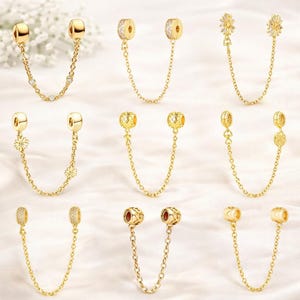 Puede incluir: Una colección de cadenas de seguridad para charms doradas. Cada cadena presenta dos cuentas de conexión con varios elementos decorativos, incluyendo cristales transparentes, diseños florales y patrones texturizados. Las cadenas están diseñadas para asegurar pulseras de abalorios.