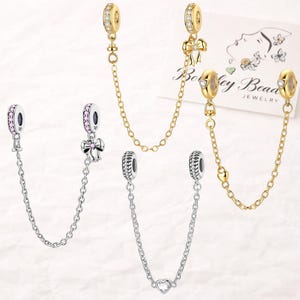 May include: Jewelry charms with safety chains. The charms are gold and silver-toned, featuring various designs like bows, hearts, and sparkling stones. The chains connect two charms, offering a secure and stylish accessory for bracelets or necklaces.