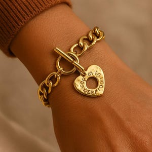 May include: Gold-coloured bracelet with a chunky chain design and a heart-shaped charm. The charm has the words "Keep me in your heart" engraved around the edge. The bracelet features a toggle clasp.