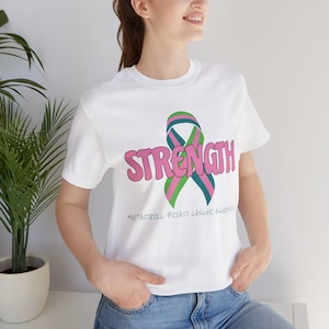 May include: A white short-sleeved t-shirt featuring the word STRENGTH in large pink bubble letters overlaid with a teal and green awareness ribbon. Below the ribbon, the text reads Metastatic Breast Cancer Awareness in a simple black font.