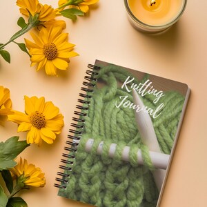 May include: A spiral-bound notebook titled "Knitting Journal" with a cover image of green yarn and knitting needles. Yellow flowers and a lit candle are arranged around the notebook on a peach-coloured surface.