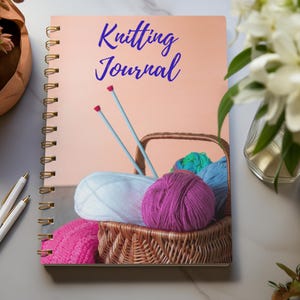 May include: A spiral-bound "Knitting Journal" with a peach cover featuring a basket of yarn and knitting needles. The journal has gold spiral binding and the words "Knitting Journal" in blue script. Two white pens are on the left.