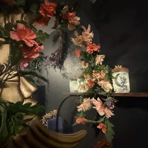 May include: A decorative display featuring artificial flowers in shades of peach and coral, arranged around a shelf. Two glass jars with a celestial design are on the shelf, illuminated by soft lighting. The scene is set against a dark wall.