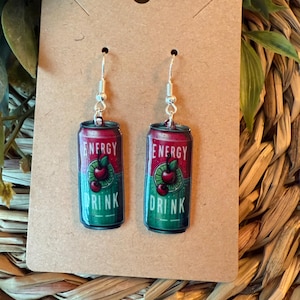 May include: A pair of dangle earrings featuring a miniature energy drink can design. The cans are red and green with the words "ENERGY DRINK" and a cherry graphic. The earrings have silver-colored fish hook ear wires.