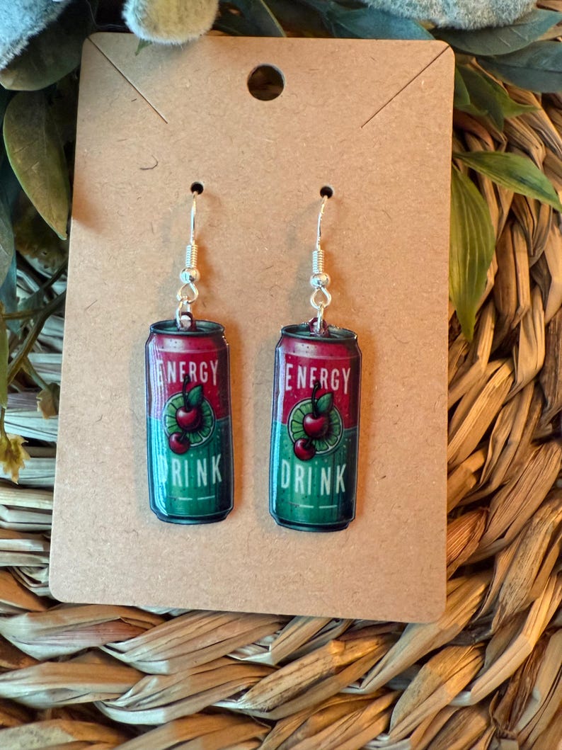 May include: A pair of dangle earrings featuring a miniature energy drink can design. The cans are red and green with the words "ENERGY DRINK" and a cherry graphic. The earrings have silver-colored fish hook ear wires.
