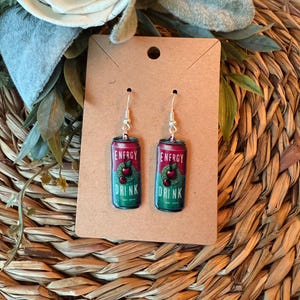 May include: A pair of novelty earrings featuring a miniature energy drink can design. The cans are red and green with the words "ENERGY DRINK" and a cherry graphic. The earrings hang from silver-colored hooks and are displayed on a brown card.