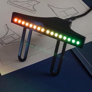 以下が含まれることがあります： カーボンファイバー調の黒いT字型電子機器。左側のオレンジ色から右側の緑色へと変化する12個のLEDライトが水平に並んで点灯しており、黒い電源ケーブルが接続されています。