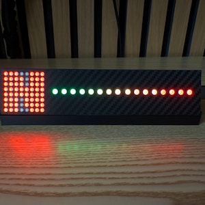 Puede incluir: Un dispositivo visualizador de audio electrónico rectangular con un acabado de textura de fibra de carbono negra. El panel frontal presenta una rejilla cuadrada de luces LED rojas a la izquierda y una fila horizontal de luces LED multicolores.