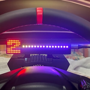 May include: A black racing steering wheel featuring a central LED display unit with a carbon fiber texture. The display shows a grid of red lights on the left and a horizontal row of bright purple lights. A cable is connected to the right side.