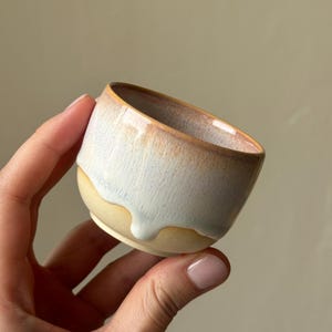 Könnte beinhalten: Eine kleine Keramiktasse mit abgerundeter Form und zweifarbiger Glasur. Der obere Teil ist in einem sanften, gesprenkelten Weiß und Hellblau gehalten, während der untere Bereich einen natürlichen beigen Ton mit sichtbaren Glasurtropfen zeigt.