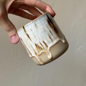 Puede incluir: Una taza de cerámica hecha a mano con forma cilíndrica. La mitad superior es blanca con goteos marrones, y la mitad inferior es de color beige. La taza está sujeta por una mano sobre un fondo neutro. La taza mide aproximadamente 9 cm de alto.