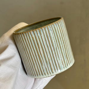 May include: A cylindrical ceramic cup featuring a light blue glaze with vertical ribbed textures. The ridges reveal the natural brown clay underneath, creating a distinct striped pattern. The interior of the cup has a smooth, muted green finish.