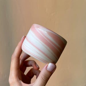 Könnte beinhalten: Ein kleiner, zylindrischer Becher mit einer glatten, glänzenden Oberfläche und einem marmorierten Muster aus sanften Pastellrosa- und Cremeweißtönen. Das Gefäß hat ein schlichtes, henkelloses Design mit abgerundetem Rand und einer klaren Optik.