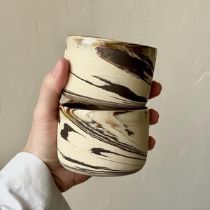 Puede incluir: Dos tazas de cerámica con base color crema y un patrón veteado de vetas marrón oscuro y negro. Las tazas son cilíndricas con un borde ligeramente ensanchado. Las tazas están apiladas una encima de la otra.