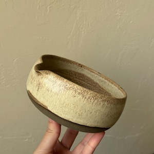 Può includere: Una ciotola in ceramica rotonda con un piccolo beccuccio. L'esterno presenta uno smalto testurizzato color crema con macchie marroni, mentre la base è di colore marrone opaco scuro. L'interno mostra uno smalto crema simile con texture argillosa.