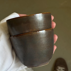 Könnte beinhalten: Zwei dunkelbraune Keramikbecher, die von einer Person gestapelt gehalten werden. Die Becher haben eine glatte, glänzende Oberfläche mit einem etwas helleren braunen Rand. Die Becher sind wahrscheinlich zum Trinken von Getränken.