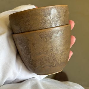 Op de afbeelding: Twee gestapelde keramische kommen met een getextureerde, metallic bronzen glazuur. De kommen hebben een licht uitlopende vorm en een gladde rand. De kommen worden waarschijnlijk gebruikt voor het serveren van voedsel of als decoratieve items.