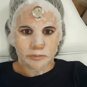 Pode incluir: Uma máscara facial branca de tecido com um dispositivo circular metálico de ligar/desligar fixado na testa. A máscara apresenta um padrão geométrico impresso e é usada com uma touca descartável branca sobre uma camisa azul-escura.