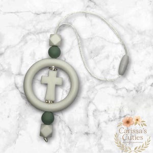 May include: A light gray teething ring with a cross design, accented with silver-colored embellishments. The ring is attached to a white cord with a gray clasp and is adorned with green and gray beads. The product is from Carissa's Cuties.