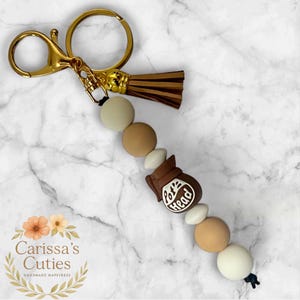 May include: A gold-colored keychain with a brown tassel and a beaded design. The beads are in shades of beige and brown, with a brown coffee pot charm that reads "Pot Head" in white. The keychain is a handmade item.
