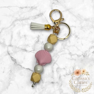 May include: A gold-toned keychain with a white tassel and a beaded strand. The strand features a pink seashell, cream-colored beads, and geometric yellow beads. The keychain is adorned with a gold clasp and ring.