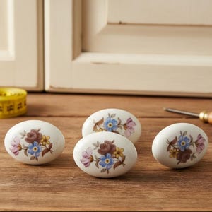 Può includere: Quattro pomelli ovali bianchi per armadi con disegni floreali blu, viola e gialli. I pomelli sono disposti su una superficie di legno. Un metro a nastro e un cacciavite sono visibili sullo sfondo.