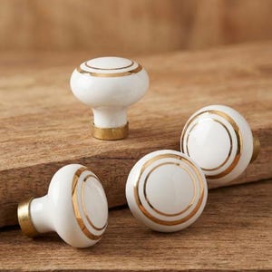 May include: Four white ceramic cabinet knobs with gold accents. The knobs have a rounded shape with a gold band around the center and a gold base. They are arranged on a wooden surface.